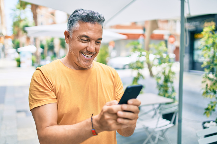 Middle age grey-haired man smiling happy using smartphone walking at street of city.