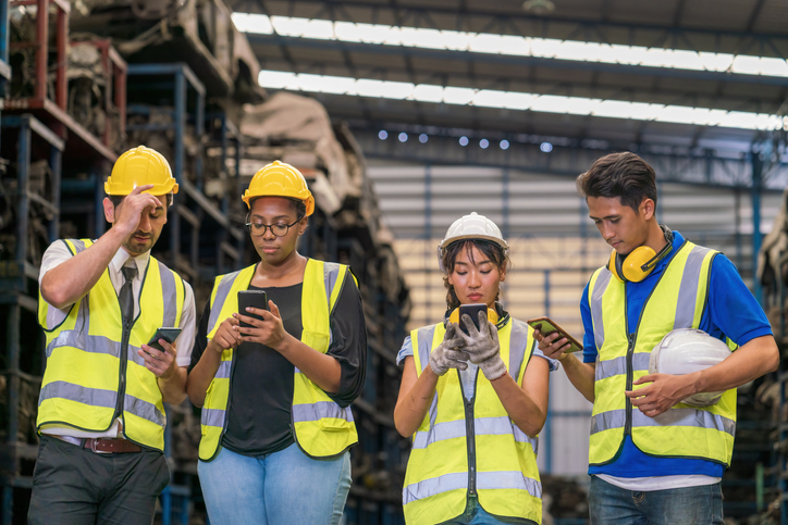 Group of manufacturer workers in hardhats and safety jackets looking at cell phones